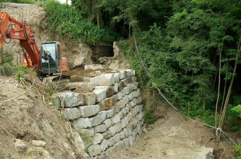 Escavatore su muro di pietra in costruzione, area boschiva.