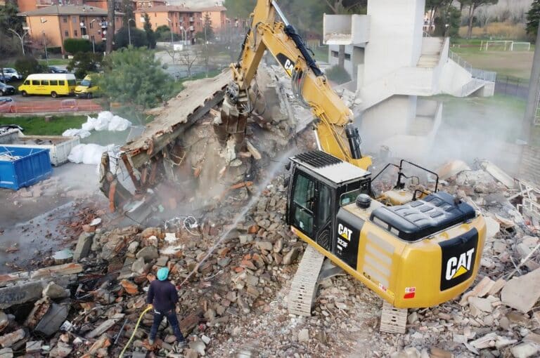 Escavatore demolisce edificio, operaio osserva tra le macerie.